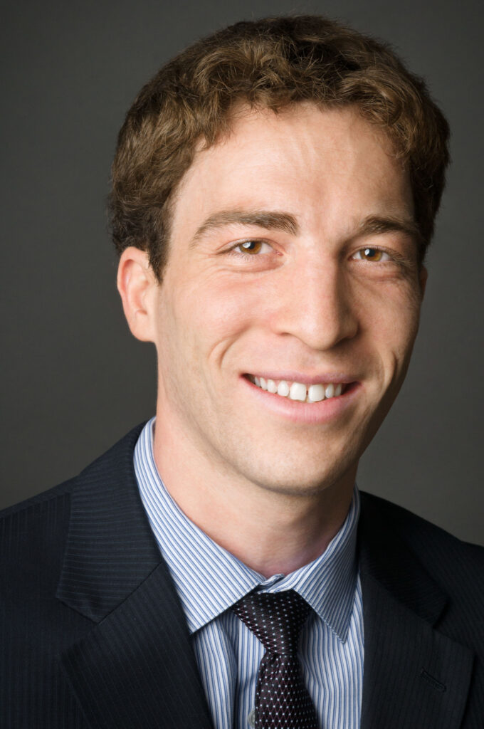 Headshot of a man in his 20s who has heritage from Brazil with light skin tone and auburn hair. He is wearing a navy pinstripe suit, white and blue striped button up shirt and navy tie. He is smiling at the camera.