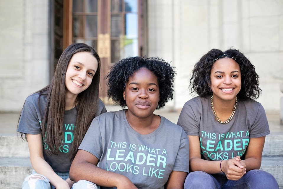 Photograph of people sitting and looking at the camera. They are all wearing the same grey t-shirt that says "This is what a leader looks like".