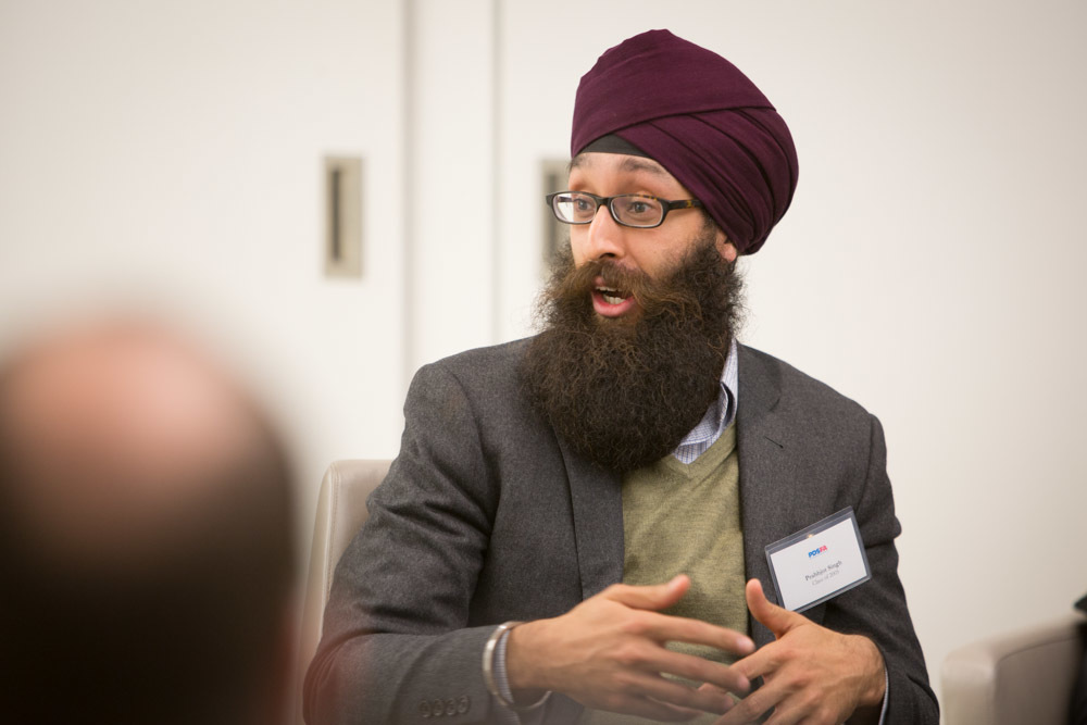 Photograph of a man in his 30s who has heritage from Kenya with light skin tone, a long dark brown beard and he is wearing a turban. He is wearing a grey blazer, olive green sweater and button up underneath, he has on oval glasses. He is looking away from the camera at someone else and talking.