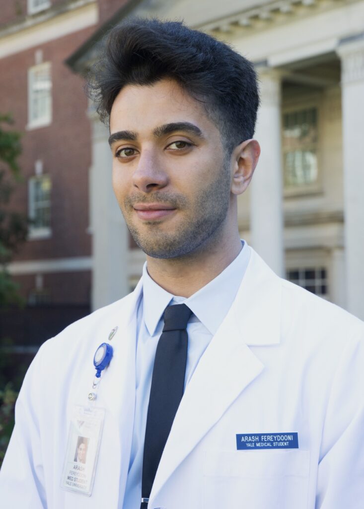 Headshot of a man in his 20s who has heritage from Iran with light skin tone and black combed back hair. He is wearing a white doctor's coat with a light blue button up and navy tie, he has a nametag on his left breast pocket and an ID badge on his right. He is smiling at the camera.