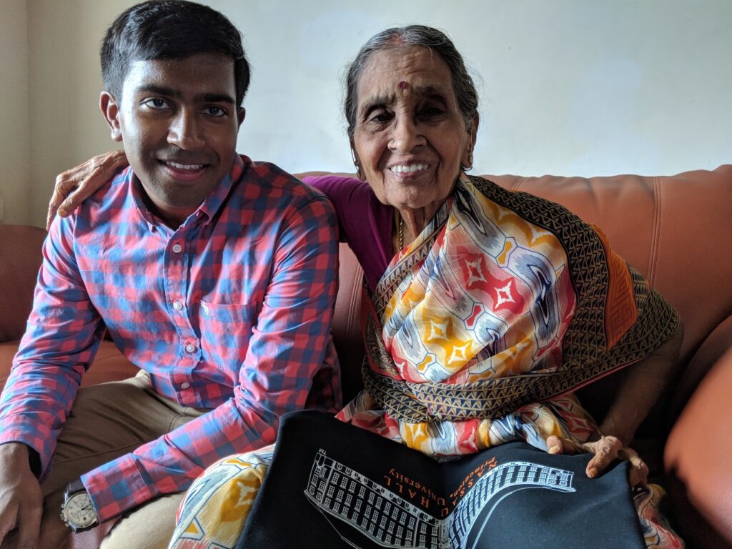 Photograph of two people sitting on an orange leather couch. The man on the left is in his 20s and wearing a blue and red gingham button up shirt and khaki pants. The woman is in her 80s and wearing a multi-colored saari, she has her arm around the man. They are both smiling at the camera. 