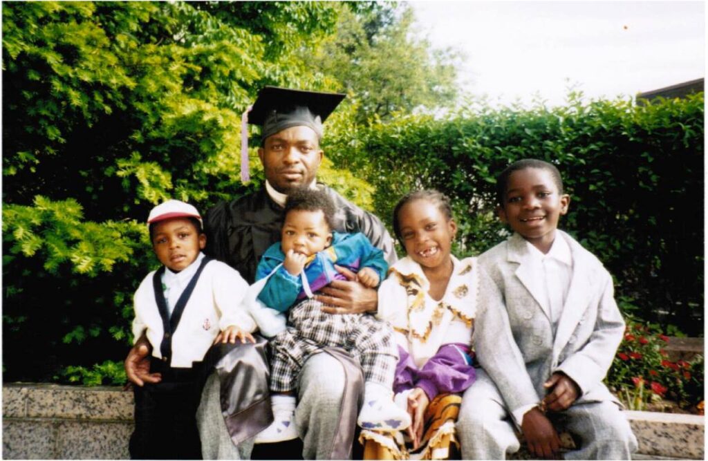 Old photograph of a man with dark skin tone and four children sitting on a ledge with trees and flowers behind them. 