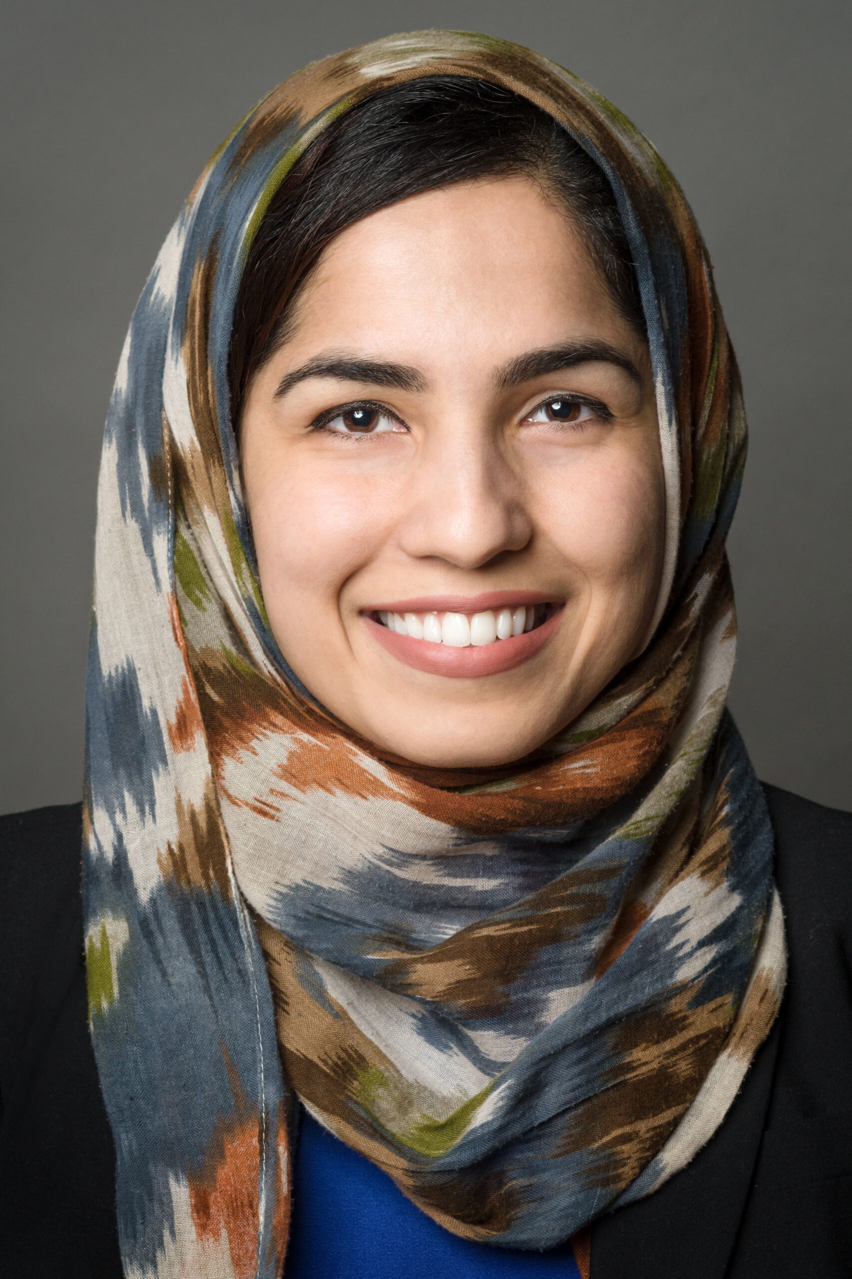 Headshot of a woman in her 20s who has heritage from Afghanistan, with light skin tone and black side bangs peeking out from a multi-colored hijab. She is also wearing a blue top and a black jacket. She is smiling at the camera.