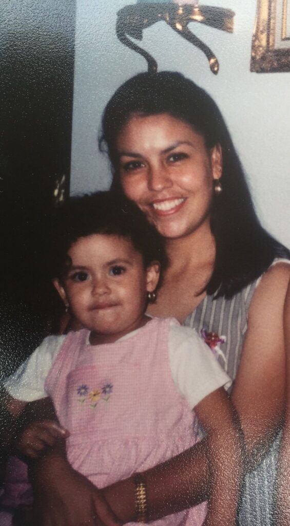 Old photograph of a woman in her 30s with light skin tone and shoulder length black hair wearing a grey sleeveless dress. She is holding a toddler with light skin tone and short black hair, wearing a pink jumper and white t-shirt. 