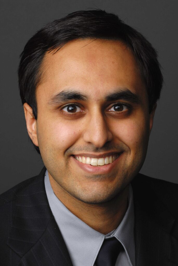 Headshot of a man in his 30s who has heritage from India with medium skin tone and black short hair; he is wearing a black pinstripe suit, grey button up shirt and black tie; he is smiling at the camera