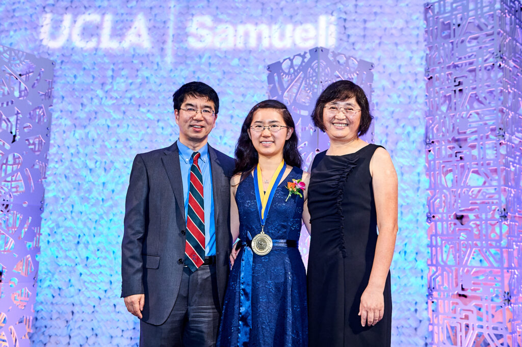 Photograph of a woman in her 20s who her Chinese heritage, she stands in the midst of white scenery with pink, purple and teal lights. She has a blue dress, boutonniere and a medal around her neck. Her parents stand on either side, they all have their arms around each other. 