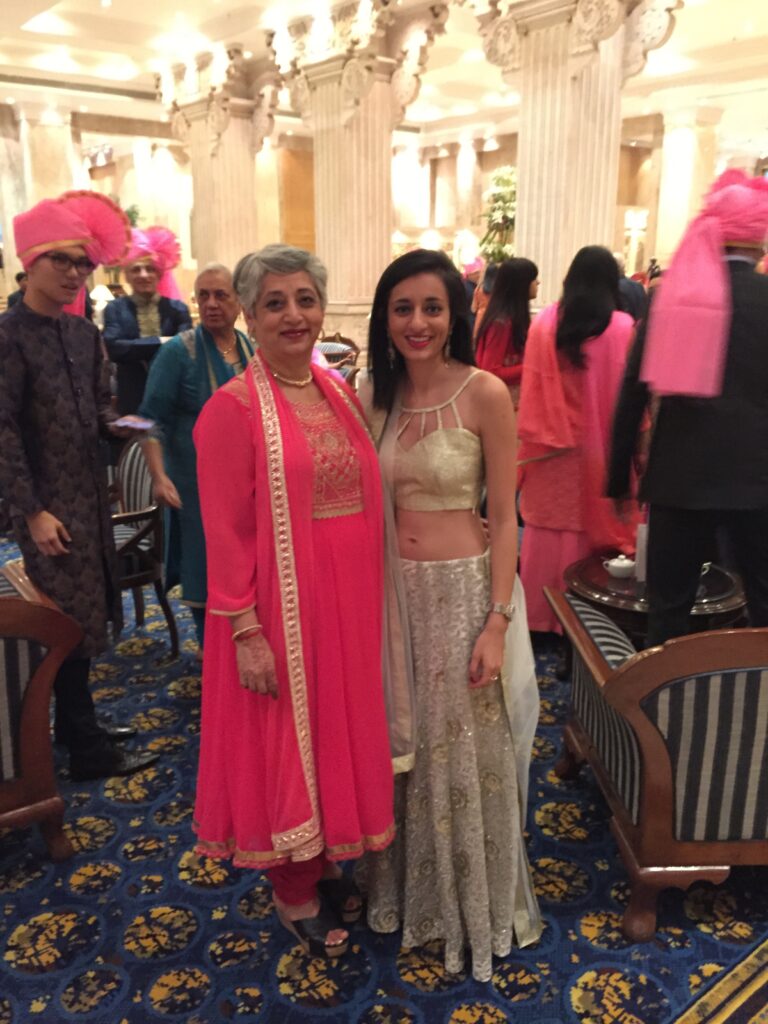 Photograph of two women standing in the middle of a party. The woman on the right is in her 20s with medium skin tone and long black hair, she is wearing a gold top and white and gold skirt. The other woman is in her 50s with medium skin tone and short grey hair, she is wearing a bright pink top and bottom. They are smiling at the camera. 