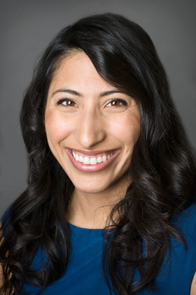 Headshot of a woman in her 20s who has heritage from Mexico with light skin tone and long curled black hair. She is wearing a blue blouse and is smiling at the camera. 