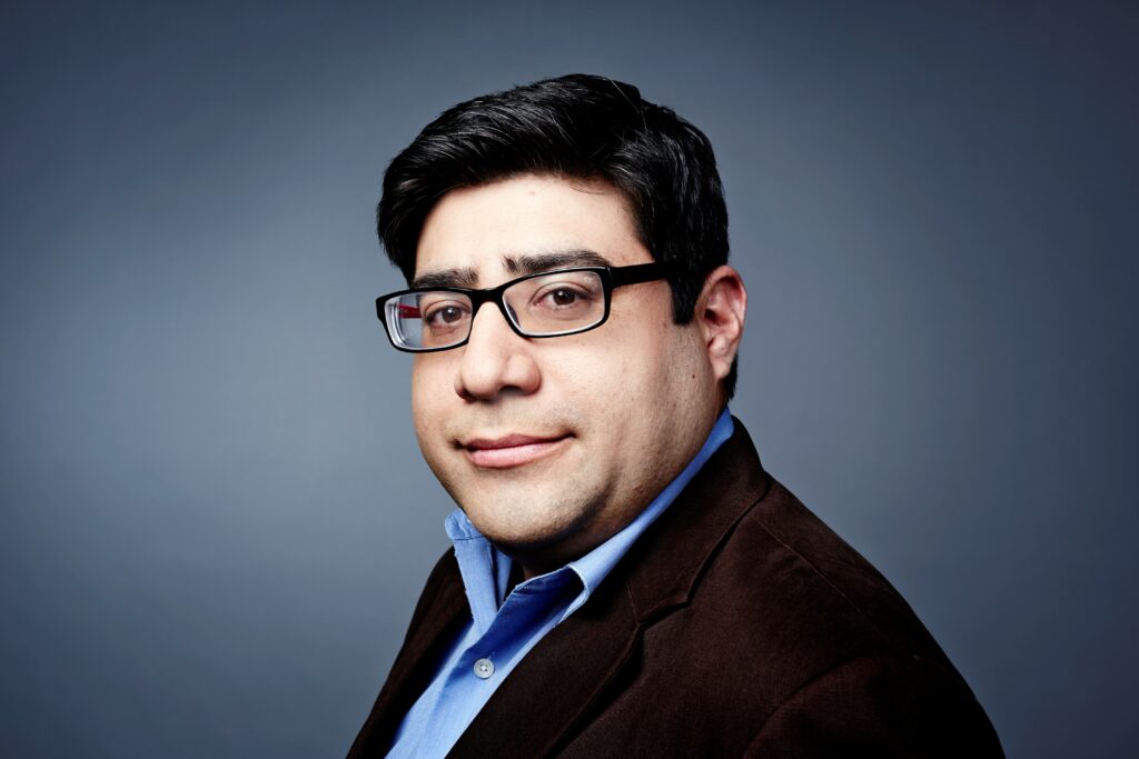 Headshot of a man in his 30s who has heritage from Peru with light skin tone and crew cut black hair. He is wearing a black suit blue button up shirt and rectangle black rimmed glasses. He is looking at the camera. 