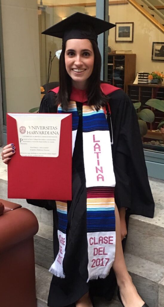 Photograph of a woman in her 20s who has heritage from Colombia, with light skin tone and long black straight hair. She is wearing black graduation robes and a grad hat with a multi-colored banner that says "Latina" around her neck. She is smiling at the camera and holding her diploma.