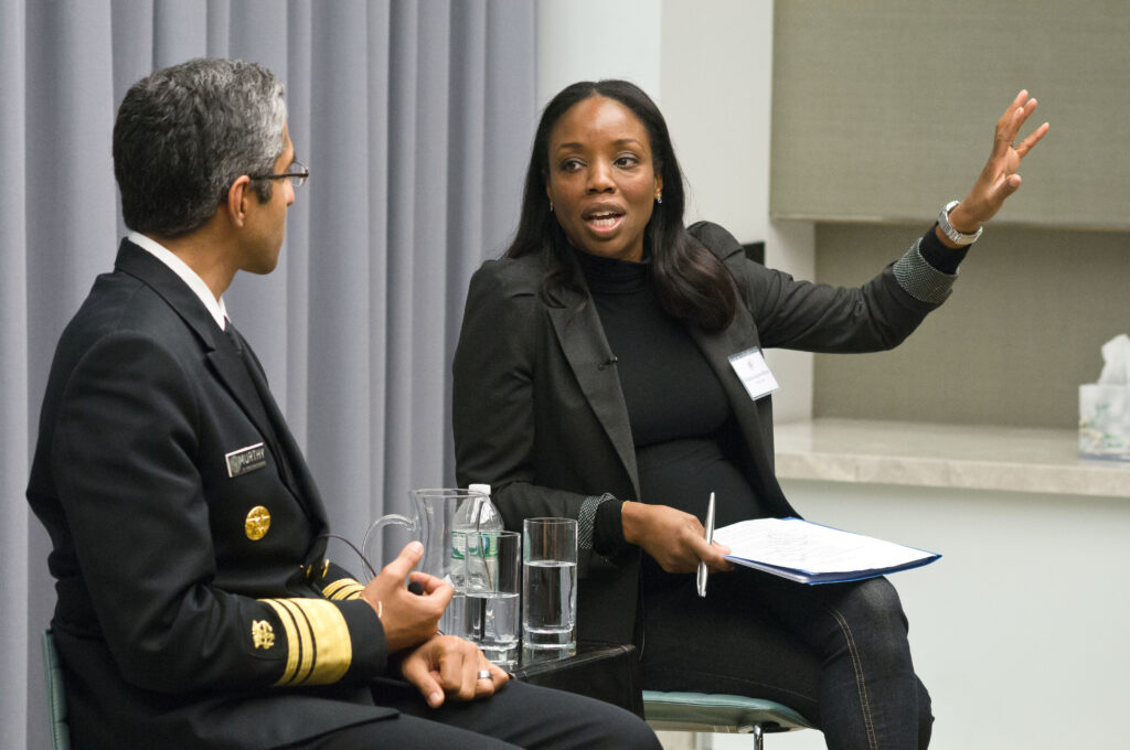 Photograph of two people sitting and talking in front of a grey curtain, the man has his back to the camera. The woman is in her 40s with heritage from Canada, she had medium-dark skin tone and long straight black hair. She is wearing a grey blazer, black turtleneck and dark jeans. 