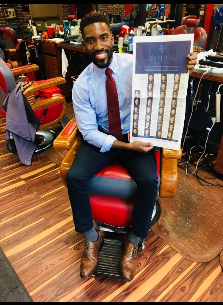 Photograph of a man in his 20s who has heritage from Nigeria with dark-medium skin tone, short black hair and a trim beard. He is w earing a light blue button up, red tied and black pants. He is holding a New York Times and seated in an orange barber chair, behind him more barber stations can be seen. 