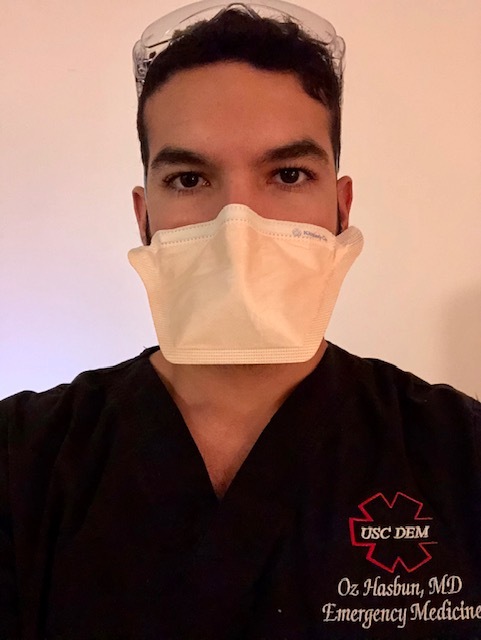 Selfie of a man in his 30s who has heritage from El Salvador with light skin tone and black hair. He is wearing black scrubs with "USC Dem / Oz Hasbun, MD / Emergency Medicine" embroidered on his left side. He has on an N95 mask and goggles on his head.