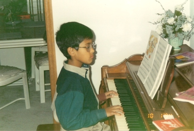 Old photograph of a young boy with medium skin tone and short black hair, wearing a blue, green and grey button up shirt and playing piano. 