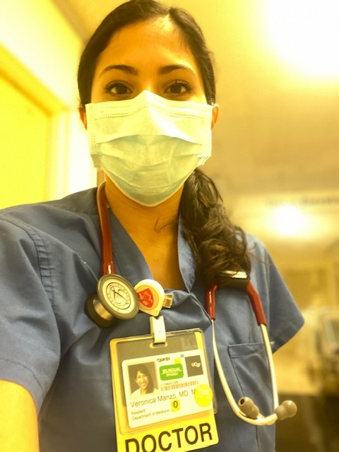 Selfie of a woman in her 20s who has heritage from Mexico with light skin tone and long curled black hair. She is wearing blue scrubs, a surgical mask, stethoscope and a "Doctor" badge.