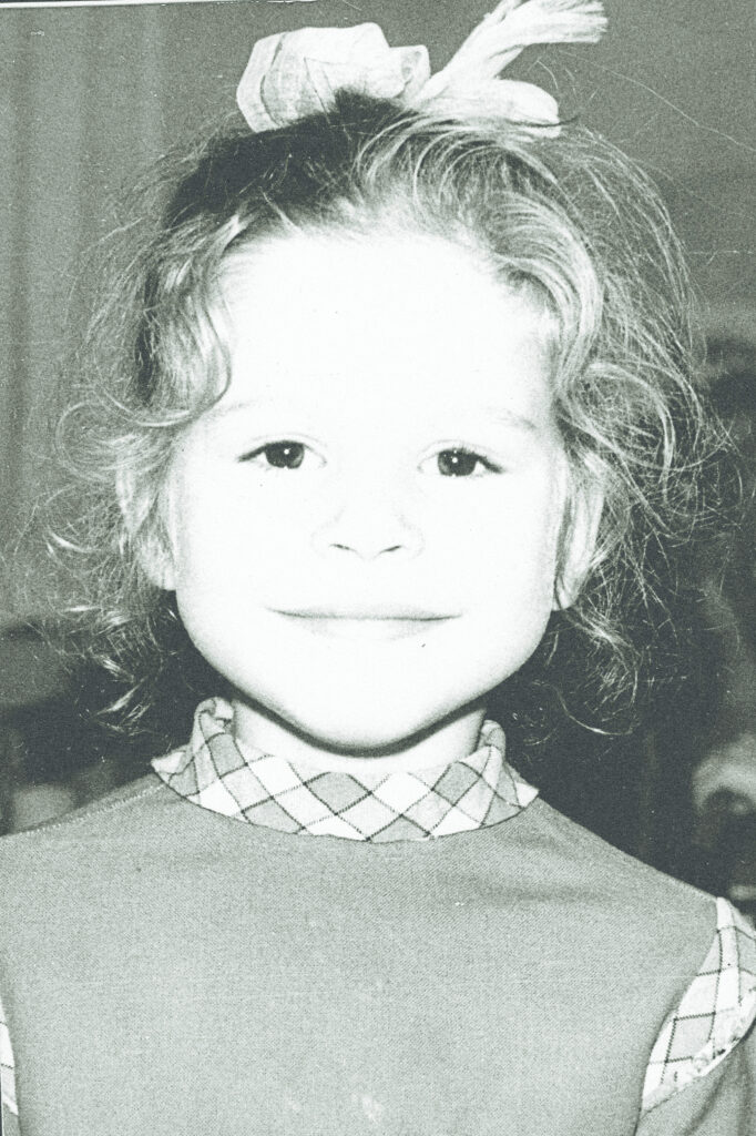 A black and white photograph of a young girl looking at the camera with a smile on her face, she is wearing a high neck top with matching accents, her hair is pulled back with a ribbon and loose curls frame her face. 