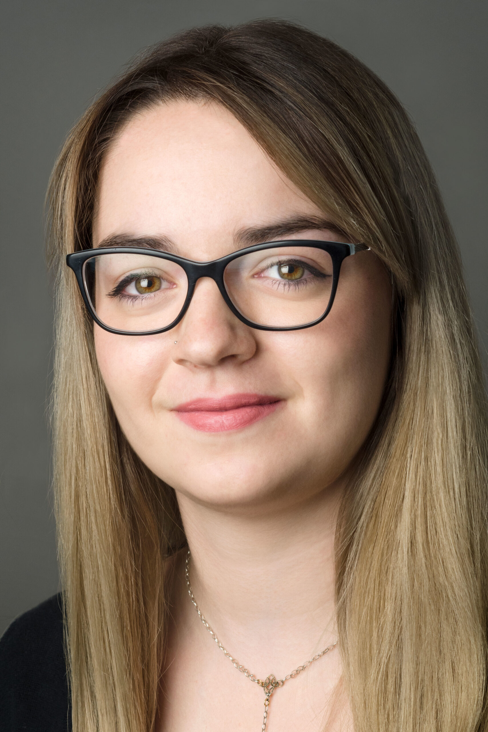 Headshot of a woman in her 20s who has heritage from Romania with light skin tone and long straight blonde hair. She is wearing a black top, gold necklace and black framed glasses. She is looking at the camera.