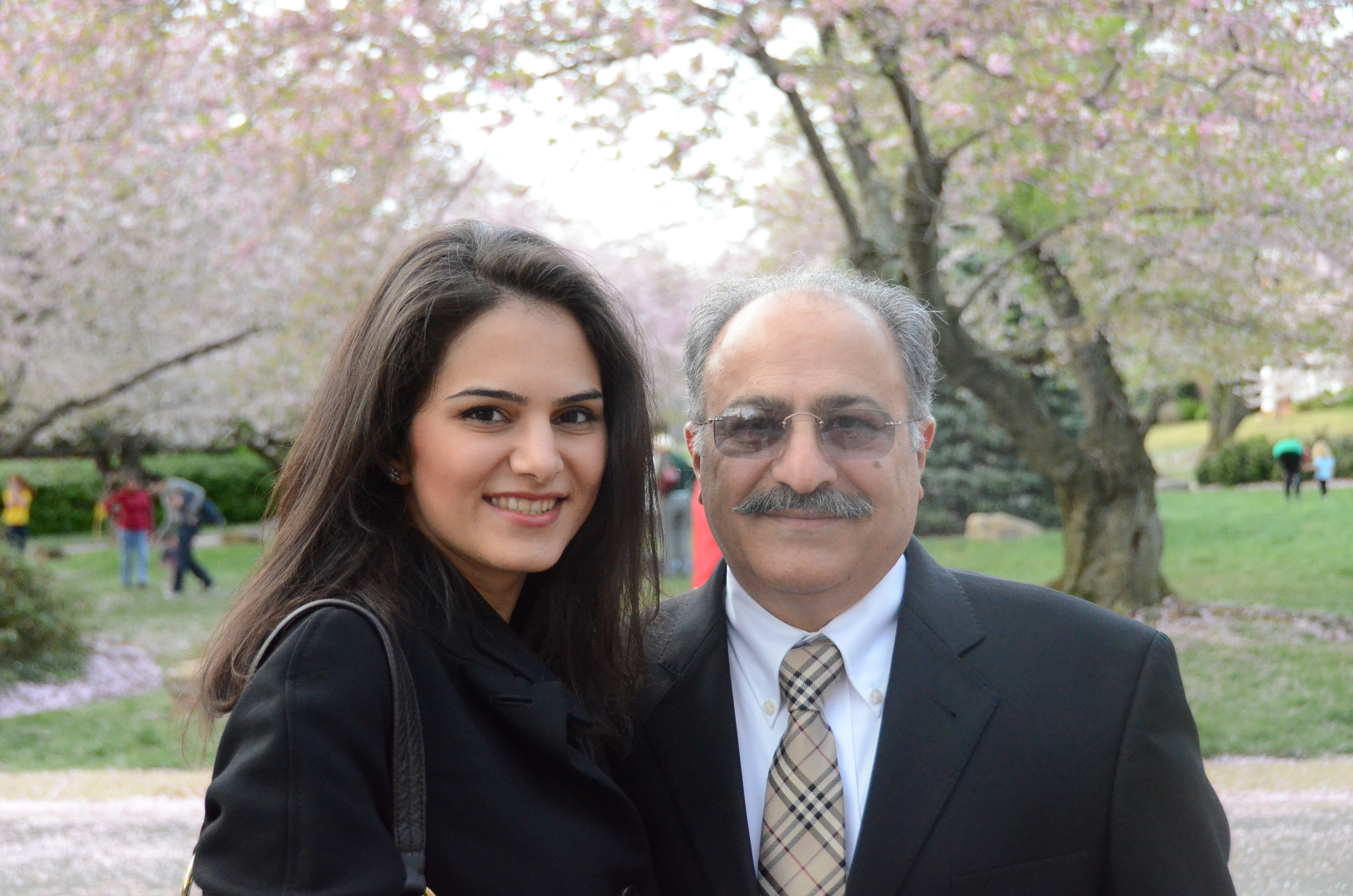 Photograph of a woman in her 20s who has heritage from Iran with light skin tone and long dark brown straight hair, she is wearing a black coat and she stands close to the man in the photo. He is in his 50s with light skin tone and grey balding hair, with a mustache. He is wearing a black suit, white button up shirt and tan tie. 