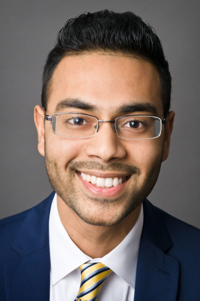 Headshot of a man in his 20s who has heritage from Bangladesh, he has medium-light skin tone with black crew cut hair and a buzzed beard. He is wearing a blue suit, white button up shirt and yellow tie, he has on frameless oval glasses. He is smiling at the camera. 