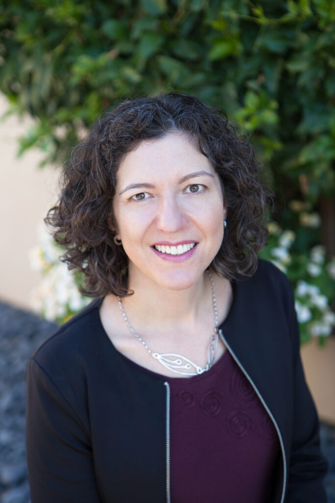 Photograph of a woman in her 30s, she has heritage from Canada, with light skin tone and chin length dark brown curly hair with visible greys. She is wearing a dark purple shirt, a black collarless jacket, a silver necklace, and small hoop earrings. She is looking slightly up at the camera and smiling. 