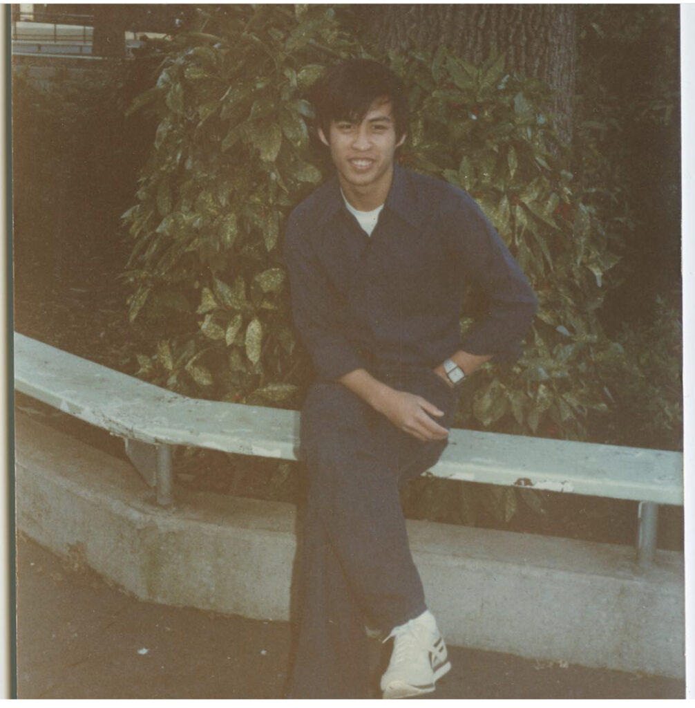 Old photograph of a young man sitting on a bench that follows a sidewalk edge, bushes and trees can be seen behind him. He is wearing a dark blue onesie. 