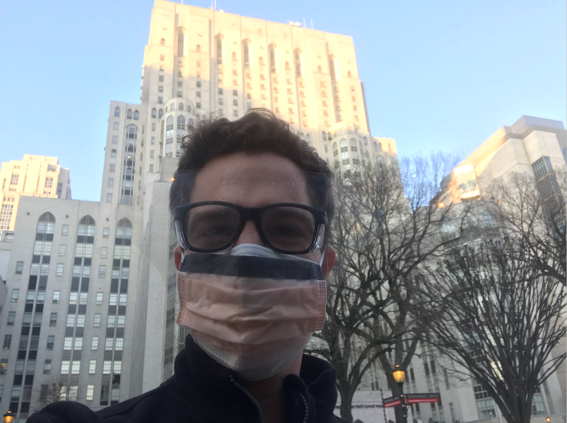 Selfie of a man in his 30s who has heritage from Brazil with light skin tone and auburn hair. He is wearing black rimmed large glasses and double surgical masks, only his black coat collar can be seen. There is a hospital building behind him.