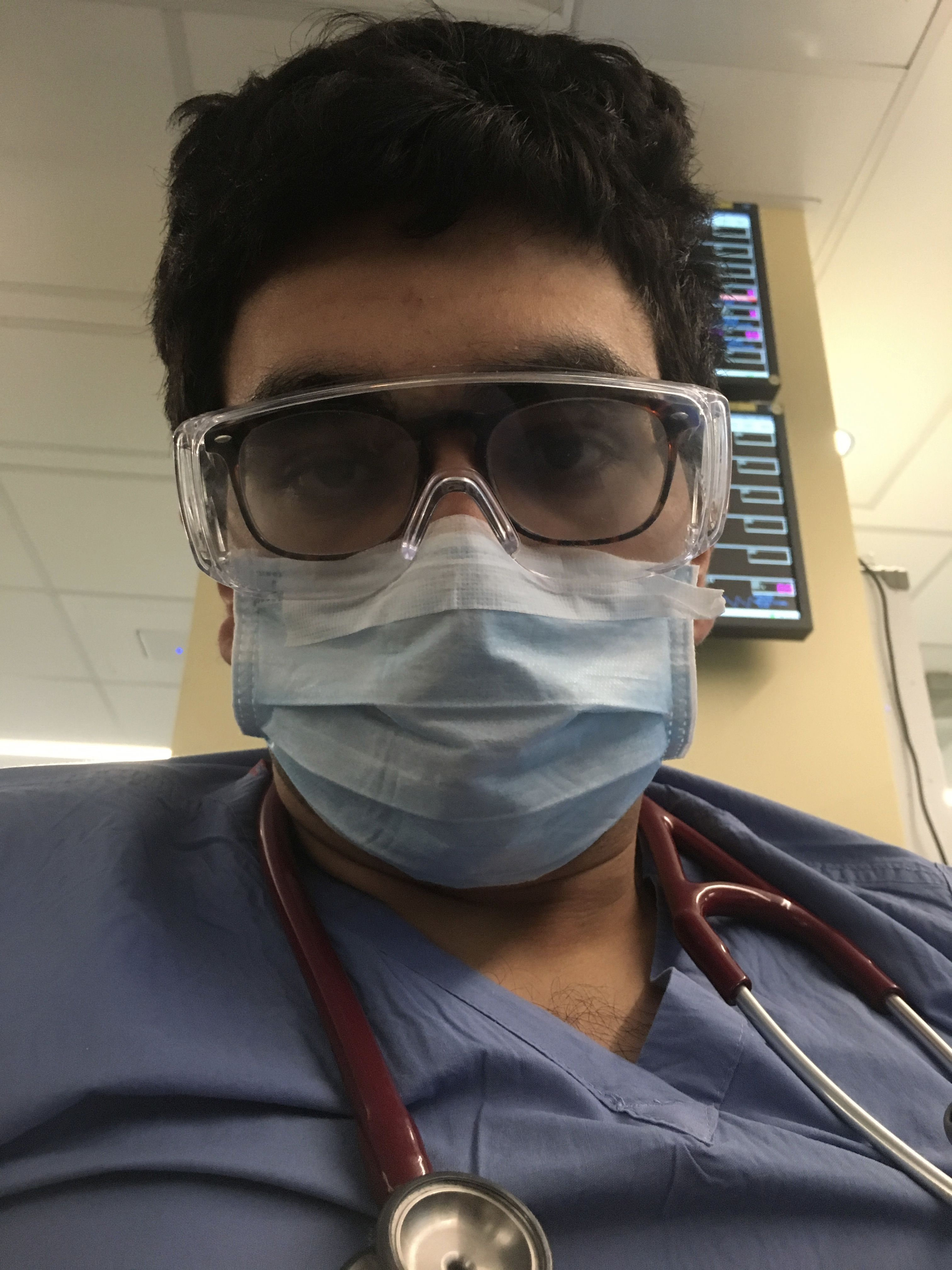 Selfie of a man in his 20s who has heritage from India with light skin tone and black hair. He is wearing blue scrubs, a surgical mask and goggles, over his glasses, as well as a stethoscope around his neck.