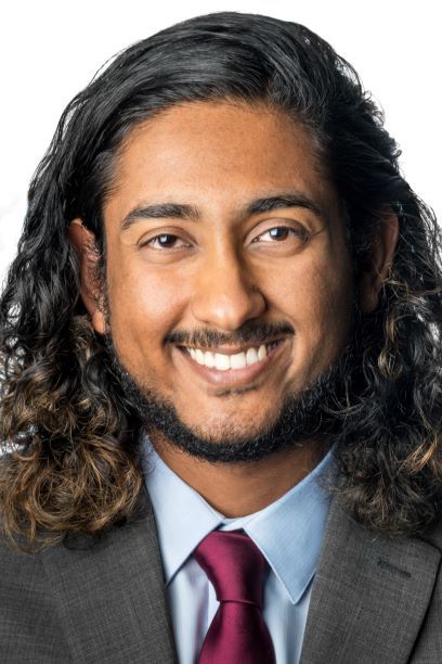 Headshot of a man in his 20s who has medium skin tone and shoulder length black curly hair. He is wearing a grey suit, light blue button up shirt and maroon tie. He is smiling at the camera.