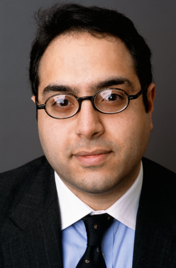 Headshot of a man in his 20s who has heritage from Egypt with light skin tone and short black hair. He is wearing a black suit, light blue with white collar button up shirt and black tie, with black thin rimmed oval glasses. He is smiling at the camera. 