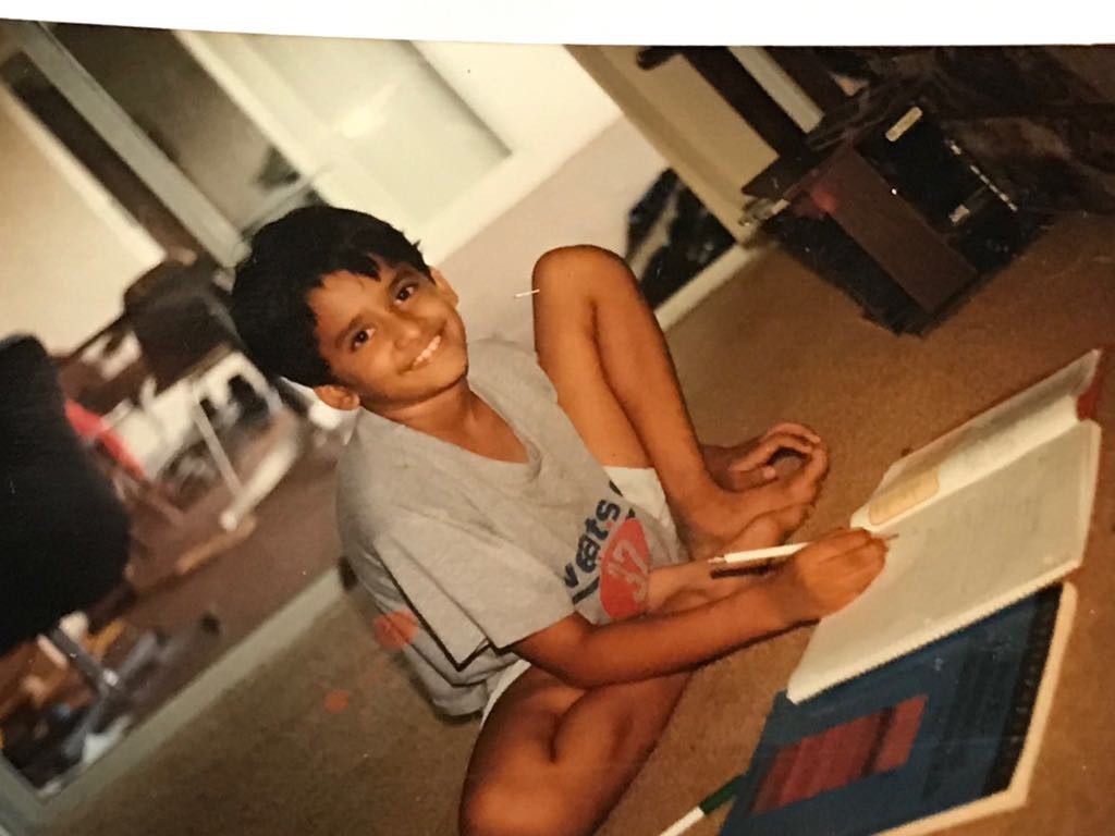 Old photograph of a young boy with medium skin tone and black hair sitting on the ground and writing in a workbook. 