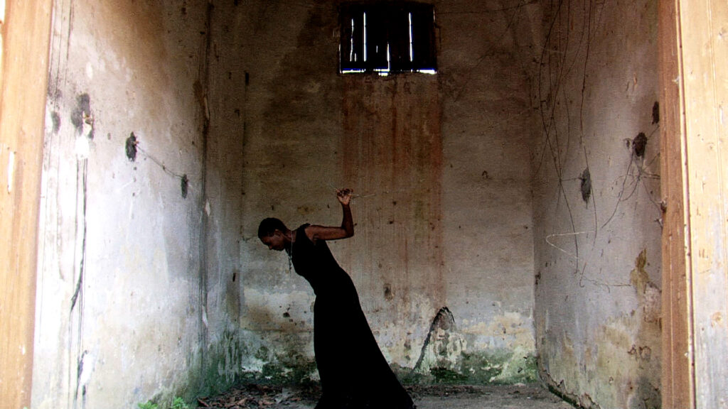 Photograph of a person in all black with dark skin tone in a room made of concrete, it has high ceilings and the appearance of rust runs down one wall. 