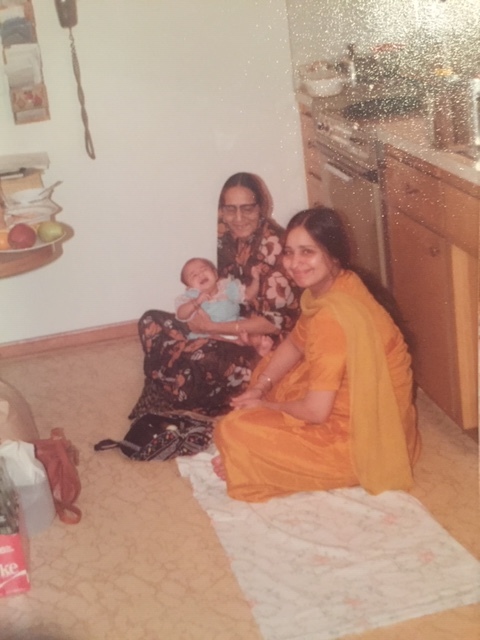 Photograph of two women and a baby. The women are dressed in sarees - one black with large pink flowers and the other orange. They are sitting on the ground in what looks to be a kitchen, the woman in black holding the baby. They are booth looking at the camera and smiling.