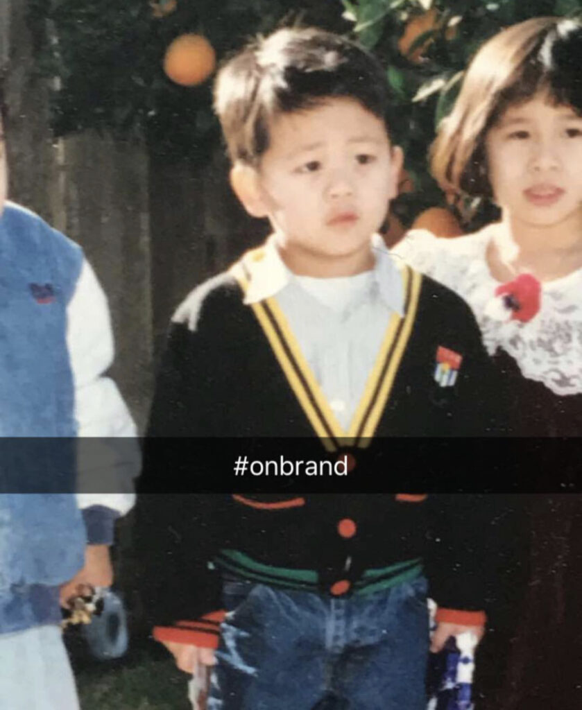 Old photograph of a young boy wearing a white button up shirt, black cardigan with yellow, red and green accents. "#onbrand" appears across the middle of the image. 