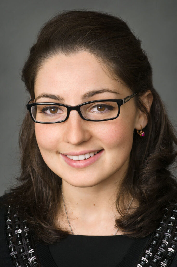 Headshot of a woman in her 20s who has heritage from Azerbaijan with light skin tone and dark brown shoulder length, half pulled back; she is wearing a black top with a sweater that has a black beaded neckline, a silver necklace, red jeweled earrings and thin black rectangle framed glasses; she is smiling at the camera