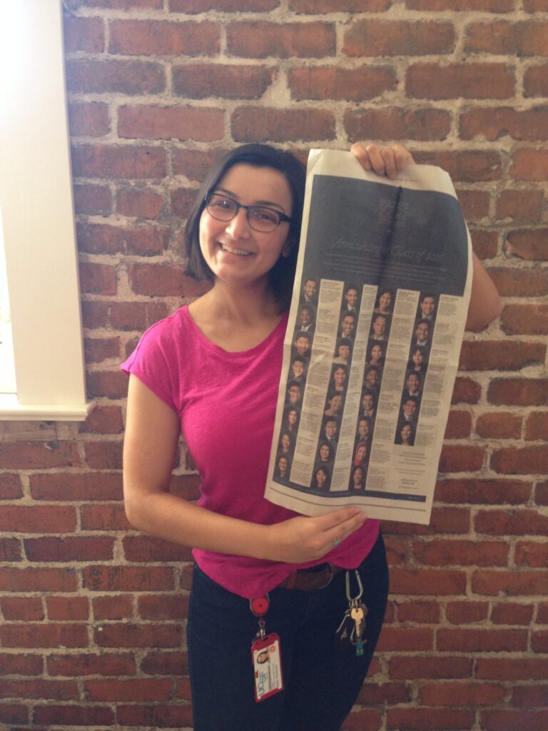 Photograph of a woman in her 20s with light skin tone and chin length black hair. She is wearing a bright pink t-shirt, and black rimmed glasses, standing in front of a red brick wall and holding up a New York Times.