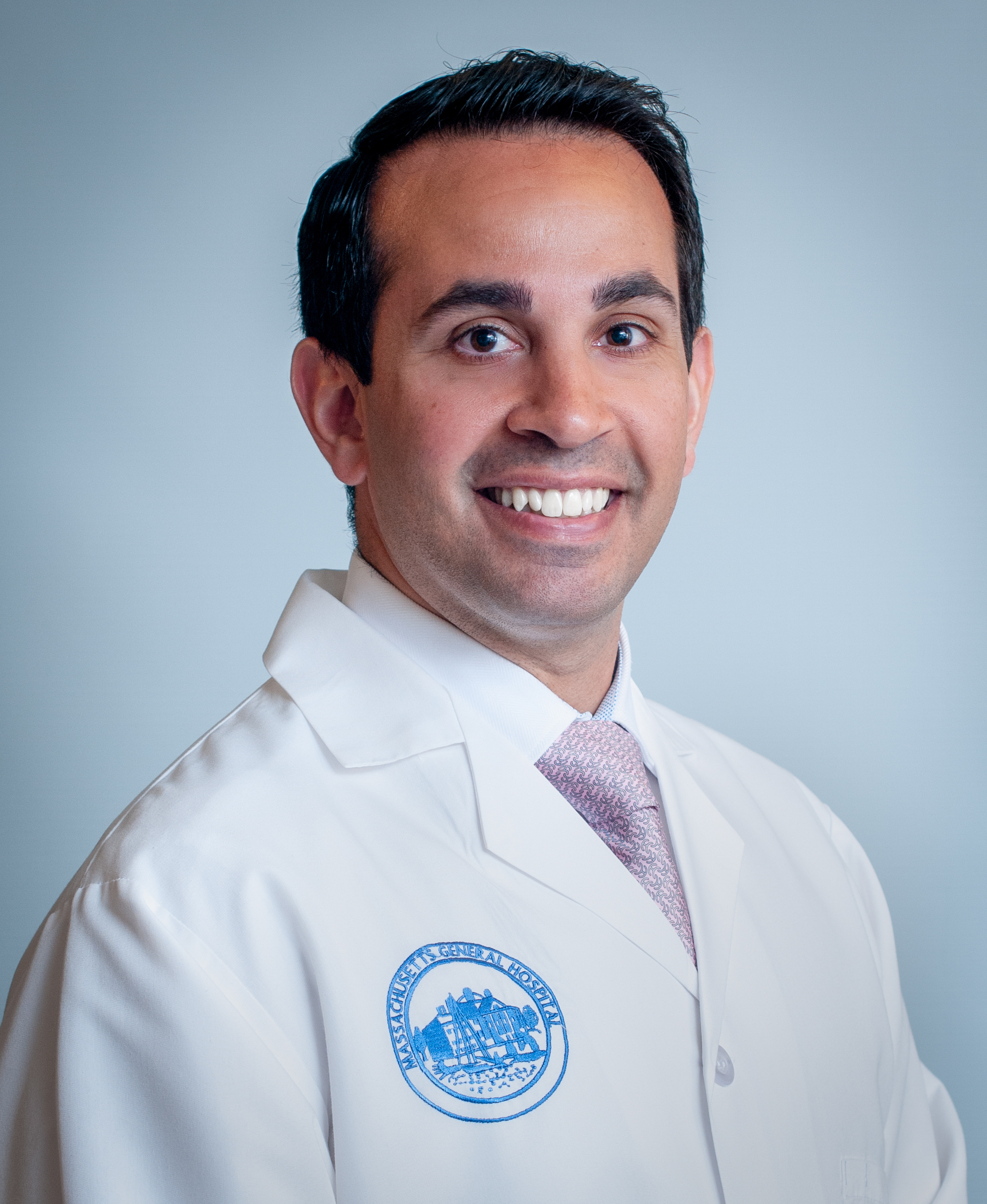 Headshot of a man in his 40s who has heritage from Iran with light-medium skin tone and black short crew cut hair. He is wearing a doctor's white coat, white button up shirt and a pink tie. He is smiling at the camera. 