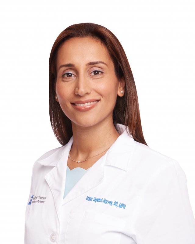 Headshot of a woman in her 30s who has heritage from Iran with medium skin tone and long straight red hair. She is wearing a doctor's white coat with a light blue blouse, gold necklace and pearl stud earrings. She is smiling at the camera.