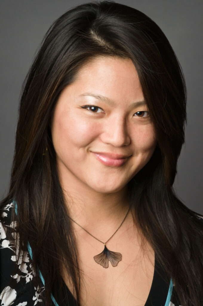 Headshot of a woman in her 20s who has heritage from China and Taiwan with light skin tone and just passed shoulder length straight black hair layered and parted on her right side. She is wearing a black and white floral-patterned top and a black gingko leaf necklace. She is smiling at the camera. 