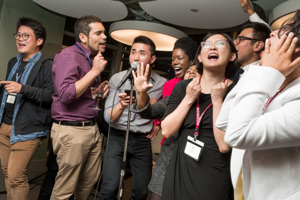 Photograph of people in their 20s with a variety of heritages standing at a microphone singing passionately. 