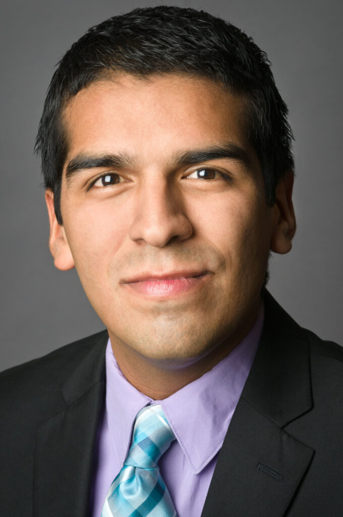 Headshot of a man in his 20s who has heritage from Mexico with medium skin tone and short black hair. He is wearing a black suit, light purple button up and blue tie. He is looking at the camera.