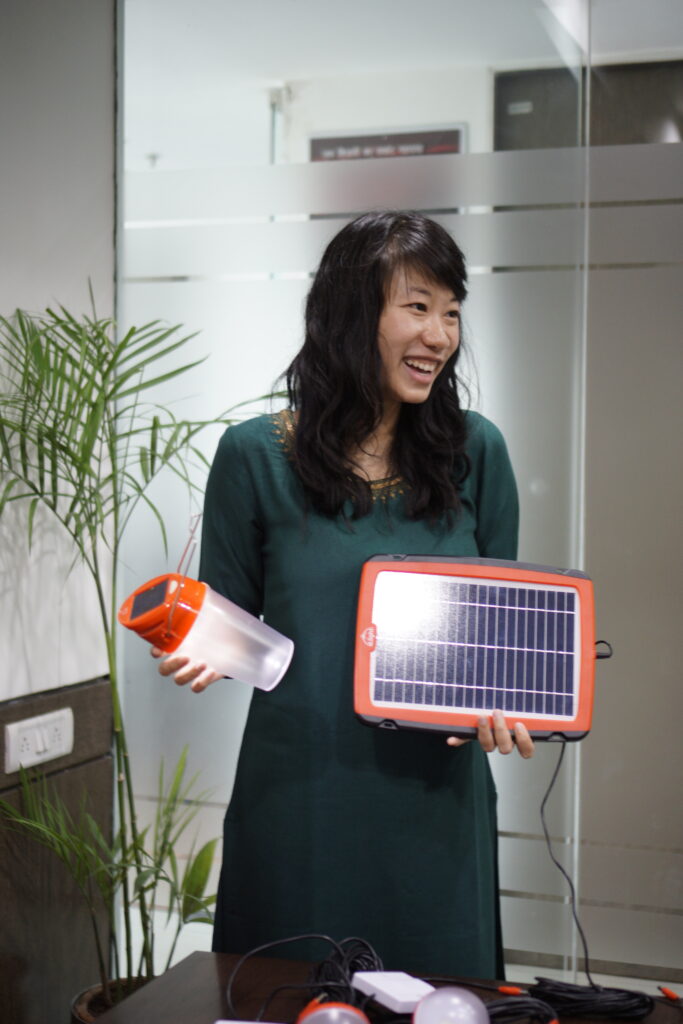 Photograph of a woman in her 20s with light skin tone and long wavy black hair, she is wearing a long dark green dress and holding a solar panel in one hand and a solar powered lantern in the other.