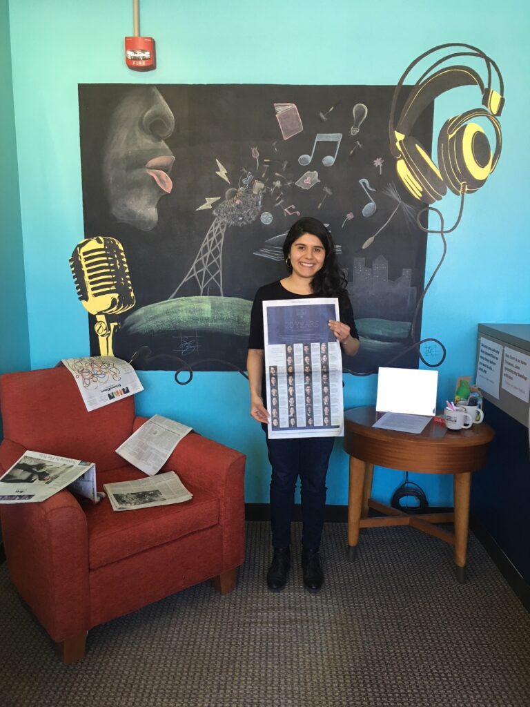 Photograph of a woman in her 20s who has heritage from Mexico with light skin tone and long black curly hair. She is wearing all black and holding a New York Times. There is a mural behind her.