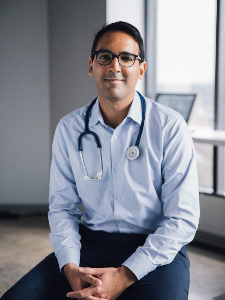 Photograph of a man sitting on a stool with windows behind him. He has heritage from Canada, medium skin tone and black short hair. He is wearing a light blue button up shirt and dark blue slacks. He has black rimmed round glasses and a stethoscope around his neck. He sits with his hands in his lap and smiling at the camera. 