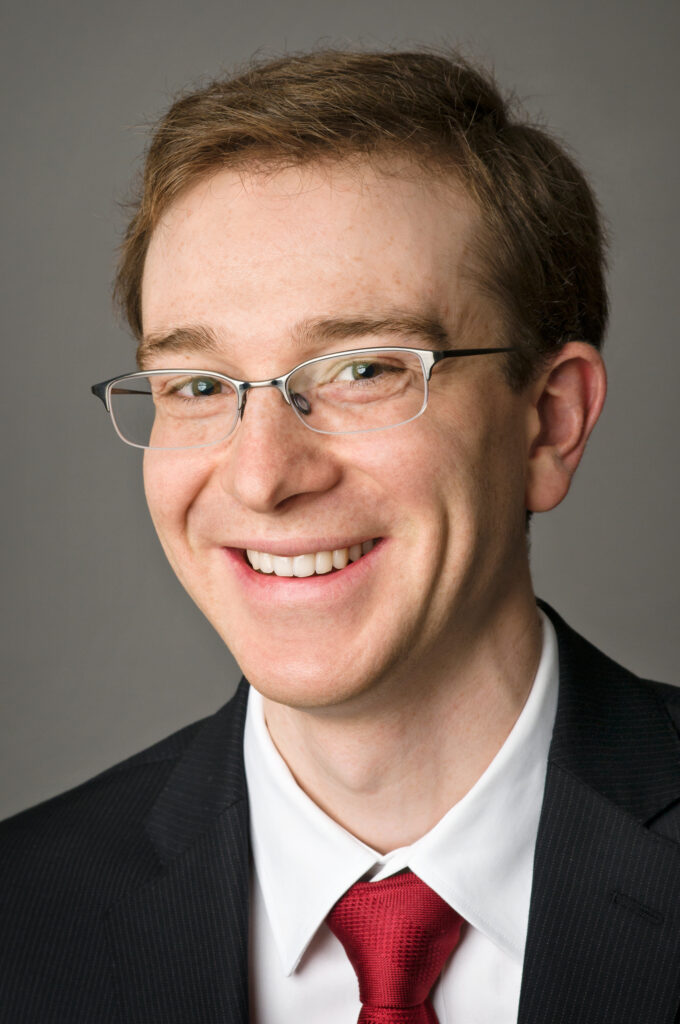 Headshot of a man in his 20s who has heritage from Ukraine with light skin tone and short auburn combed over hair. He is wearing a black pinstripe suit, white button up shirt and red tie, he has on half rimmed glasses. He is smiling at the camera. 