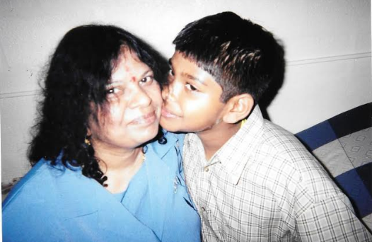 Old photograph of a woman in her 30s with light-medium skin tone and black shoulder length hair, wearing a blue top, looking at the camera while a young boy kisses her on the cheek. 