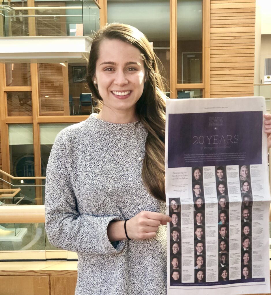 Photograph of a woman in her 20s who has heritage from Istanbul with light skin tone and long straight light brown hair. She is wearing a grey sweater and holding a New York Times. There is a building made of wood and glass behind her. 