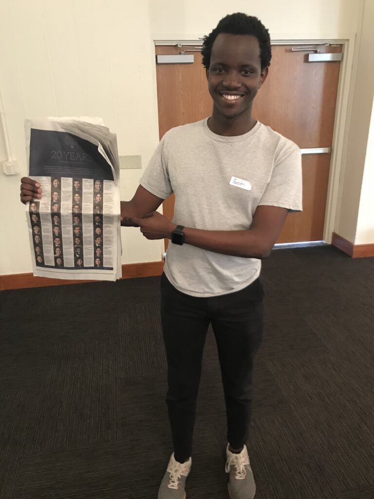 Photograph of a man in his 20s who has heritage from Kenya with dark skin tone and short black hair. He is wearing a white t-shirt and jeans, holding a New York Times. He is standing in front of a white wall and wood doors. 