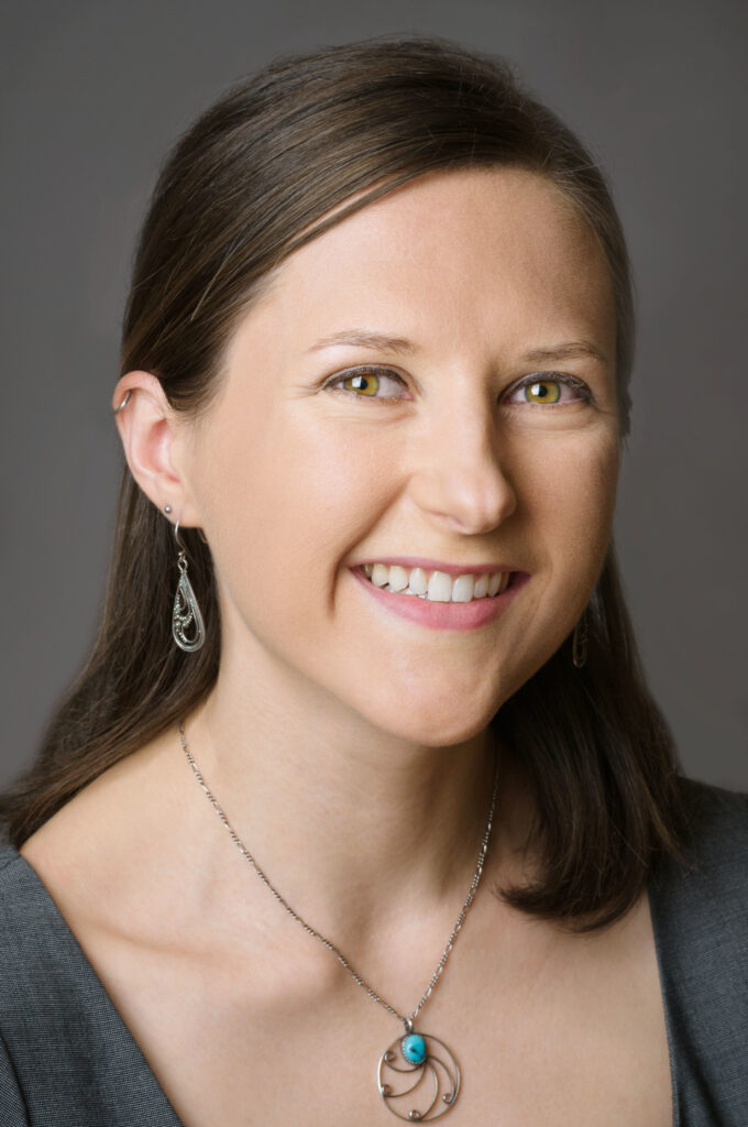 Headshot of a woman in her 20s who has heritage from Poland, she has light skin tone and shoulder length straight brown hair, half pulled back. She is wearing a grey top, silver and turquoise necklace and dangly silver earrings. She is smiling at the camera. 