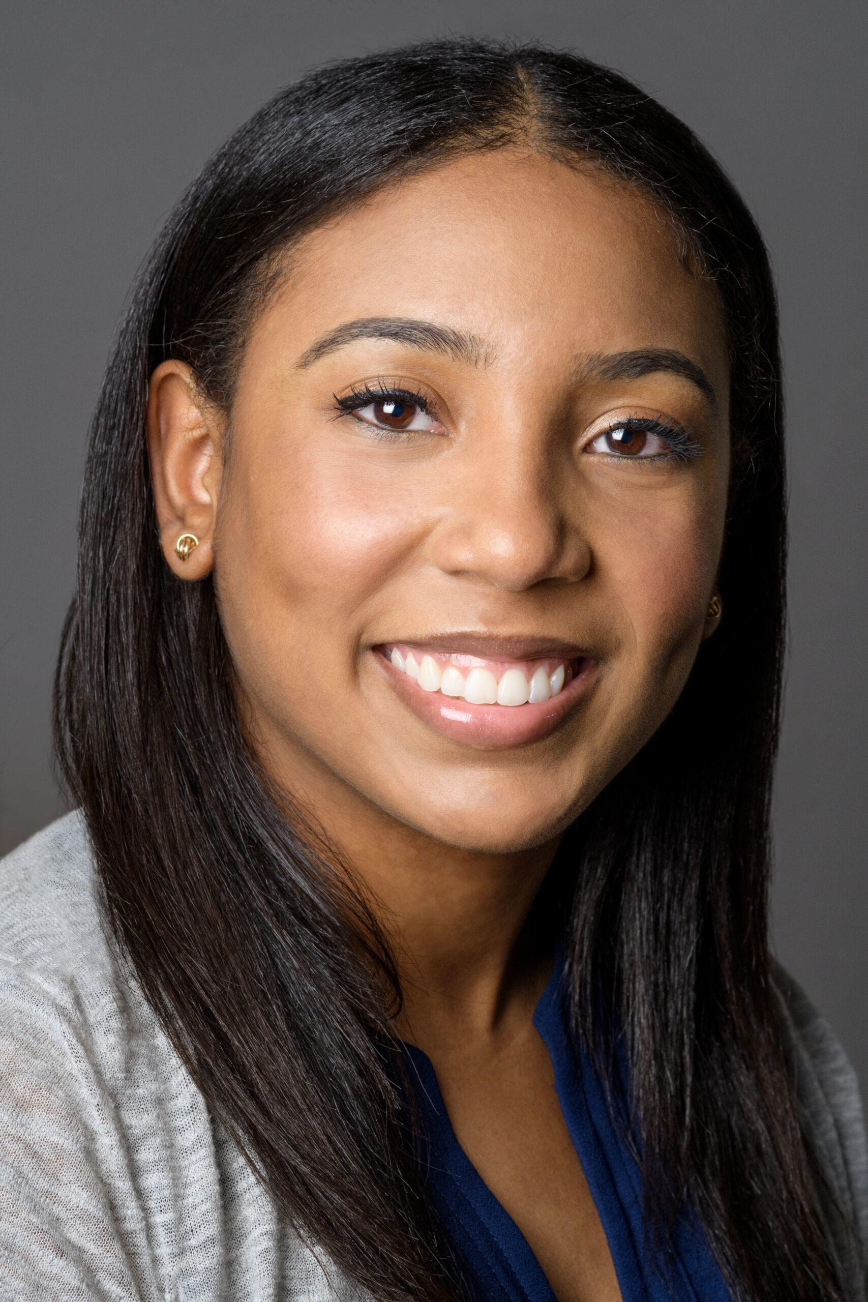 Headshot of a woman in her 20s with heritage from Dominican Republic, she has medium skin tone and straight, long black hair. She is wearing a light grey sweater, blue shirt and smiling at the camera.