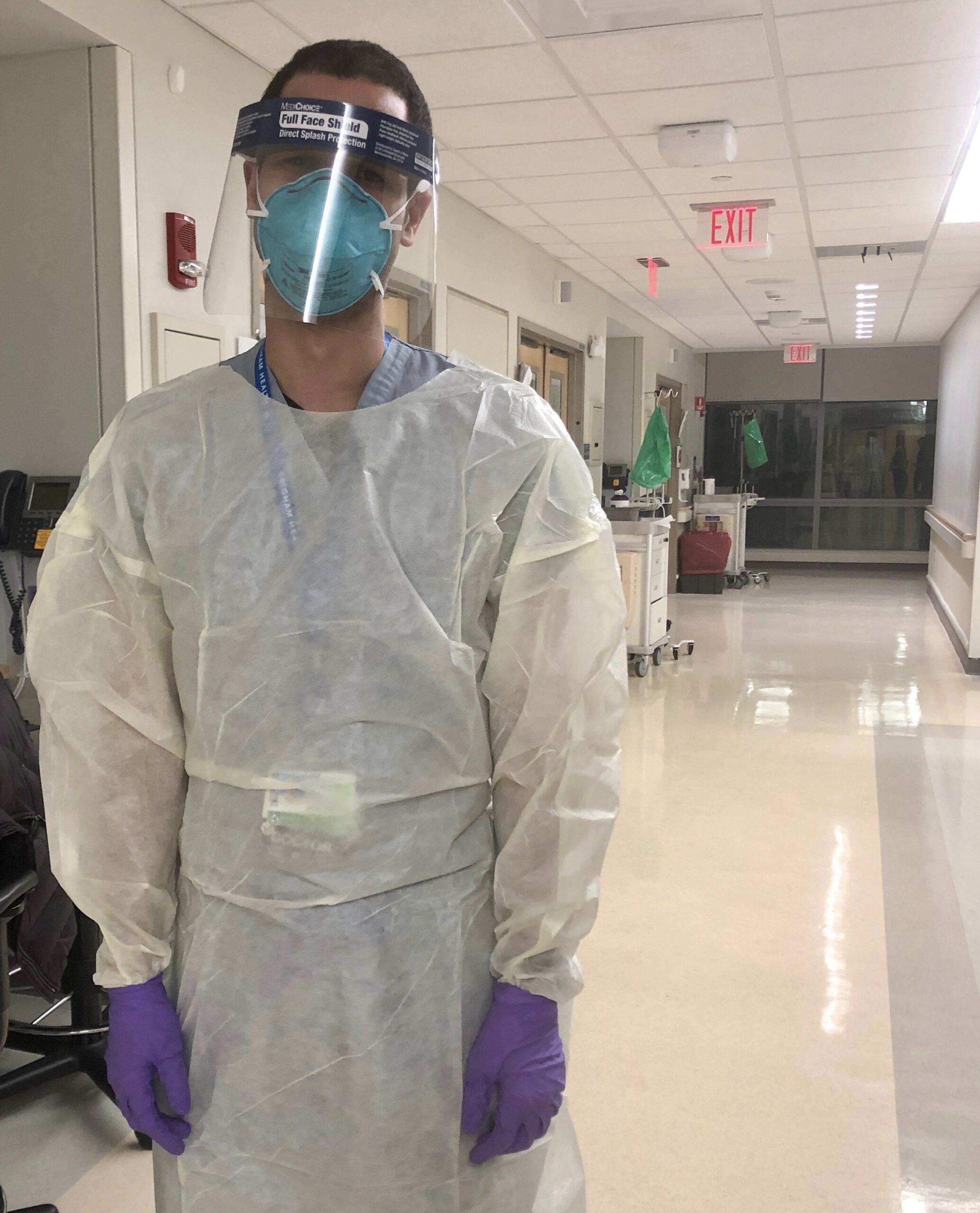 Photograph of a man in his 30s who has heritage from the Dominican Republic with light-medium skin tone and dark brown buzzed hair. He is wearing blue scrubs under full PPE (face shield, face mask, overall, gloves). He is standing in the hallway of a hospital.